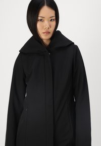 Veste à capuche noire en tissu doux. Présente un col haut, une silhouette élégante et des poches latérales. Design minimaliste sans embellissements.