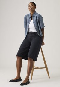 Γυναίκα με κοντά σκούρα μαλλιά, φορούσα μπλε καρό κοντομάνικο πουκάμισο πάνω από λευκή φα νέλα, μαύρα μακριά σορτς και μαύρα φλατ παπούτσια, καθισμένη σε ξύλινο σκαμπό.