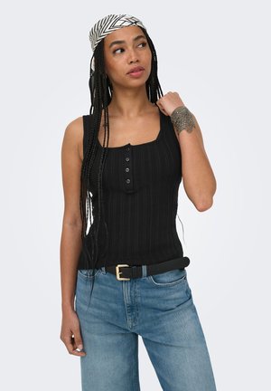Kvinde med lange fletninger iført en sort ærmeløs top, blå jeans, sort bælte, mønstret bandana og metal armring, der kigger til siden.