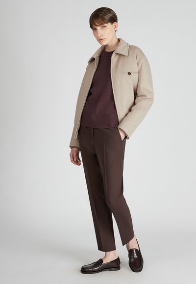 Donna in piedi con giacca beige, maglione marrone scuro, pantaloni abbinati e mocassini marrone scuro davanti a uno sfondo chiaro e uniforme.