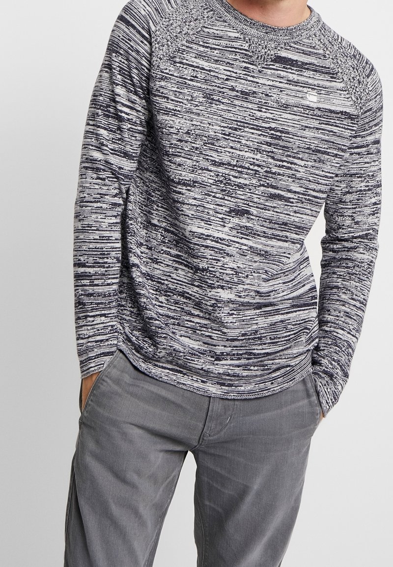 Pull à manches longues et col rond en maille chinée bleu marine et blanc. Il présente des manches raglan et un col texturé. Tissu doux.