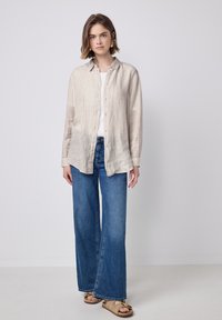 Camisa de botones beige a rayas con un corte relajado, combinada con jeans de mezclilla azul de pierna ancha. El modelo lleva sandalias neutras y una camiseta sin mangas blanca.