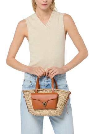 Donna con top beige senza maniche e jeans azzurri chiaro che tiene una borsa intrecciata di paglia con tasca in pelle marrone e manici.