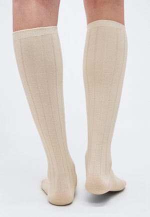 Nogi w beżowych, prążkowanych podkolanówkach sięgających do kolan, stojące na gładkiej, jasnoszarej powierzchni, widziane od tyłu.