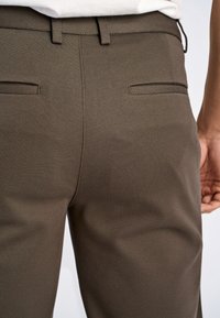 Bruine, op maat gemaakte broek met een gladde textuur, achterzakken en een aansluitende tailleband. De stof lijkt licht rekbaar.