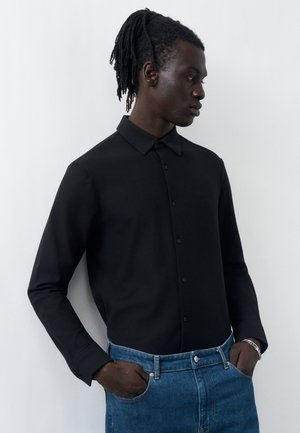 Άνδρας με ντραντλοκς που φοράει μαύρο πουκάμισο με κουμπιά και μπλε τζιν, στέκεται με τα χέρια στις τσέπες μπροστά σε απλό ανοιχτό φόντο.