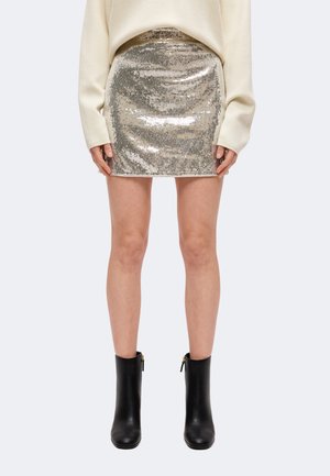 Modèle portant une minijupe argentée à sequins, un pull couleur crème et des bottines noires sur un fond uni.