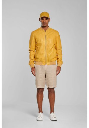 Mężczyzna w musztardowej czapce i kurtce na suwak, beżowych szortach oraz białych trampkach, stojący na jednolitym szarym tle.