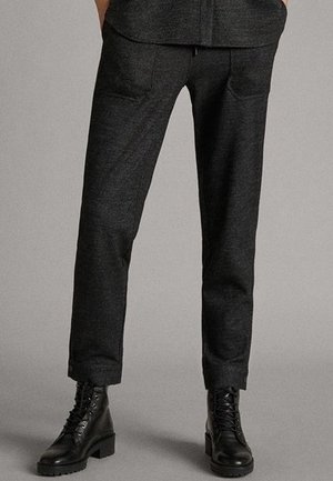 Person iført mørkegrå skræddersyede bukser og sorte snørebåndsankelstøvler, der står mod en ensfarvet grå baggrund.