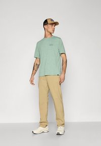 Lys grønn kortermet skjorte med et diskret logo, kombinert med beige bukser. Modellen har på seg en beige caps og hvite joggesko.