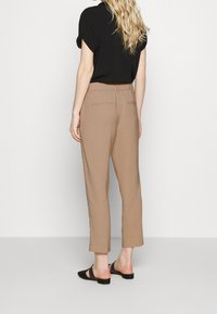 Mujer con cabello largo y rubio que lleva una blusa negra de manga corta, pantalones beiges de tiro alto y zapatos de tacón negro tipo slingback, de espaldas.