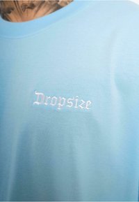 Tricou din bumbac de culoare bleu deschis, cu text brodat alb "Dropsize". Materialul este neted, cu decolteu rotund și mâneci scurte.