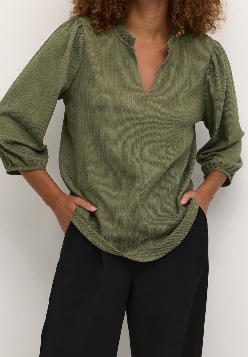 Blouse verte olive texturée avec un léger décolleté en V, manches bouffantes et ourlet incurvé. Associée à un pantalon noir.