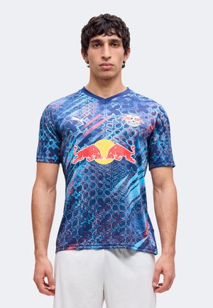 Hombre con camiseta de fútbol azul estampada del RB Leipzig con el logo de Red Bulls y pantalones cortos blancos, de pie contra un fondo claro y liso.
