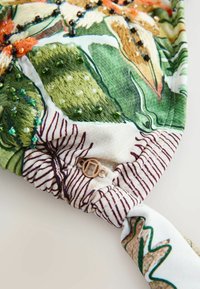 Textil fejpánt zöld levél hímzéssel, több színű gyöngyökkel és egy kicsi, kerek arany fém medállal, fehér és csíkos háttéren.