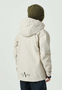 Beige waterproof jacket with a hood, featuring elastic cuffs, side pockets, and a prominent logo on the back. Worn with a dark green knit hat.