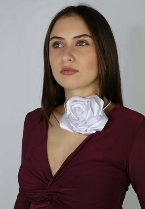 Mujer con cabello castaño lacio, lleva una blusa burdeos y un collar grande de tela blanca con forma de flor, mirando ligeramente hacia arriba y a la derecha.