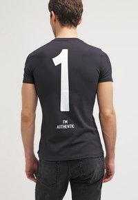 Musta lyhythihainen t-paita, jossa on selässä suuri valkoinen numero "1" ja tekstinä "I'M AUTHENTIC" numeron alla. Pehmeää materiaalia.