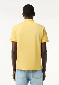 Camisa polo amarilla de manga corta y cuello, hecha de tela suave, combinada con jeans azul claro, vista desde la parte de atrás.