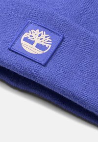 Blå stickad mössa i mjukt material med en fyrkantig patch som har en trädlogotyp i ljusbeige. Slät yta, vikbar kantdesign.