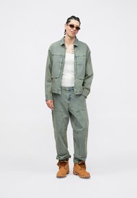 Zestaw z kurtką i spodniami jeansowymi w jasnoniebieskim kolorze o luźnym kroju; posiada głębokie kieszenie i gładką fakturę. Noszony z białą koszulką na ramiączkach i brązowymi butami.