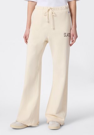 Kremowe szerokie spodnie dresowe z rozciąganym pasem i sznurkiem oraz małym napisem "S. ATL" na górnej lewej części uda, noszone z białymi tenisówkami.