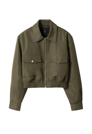 Person iført en cropped olivengrøn jakke, grøn mønstret top, høj taljede blå jeans og sorte støvler, står med hænderne på hofterne.