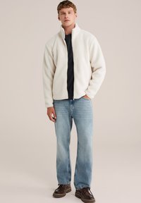 Giacca in pile color crema chiaro con colletto alto, zip frontale e maniche lunghe, abbinata a jeans blu chiaro e scarpe marrone scuro.