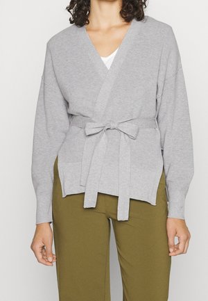 Femme portant un pull en maille gris clair avec une ceinture nouée par-dessus un haut blanc et un pantalon vert olive, visible du cou aux hanches.