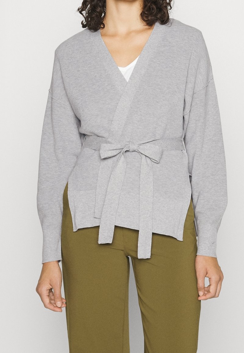 Femme portant un pull en maille gris clair avec une ceinture nouée par-dessus un haut blanc et un pantalon vert olive, visible du cou aux hanches.