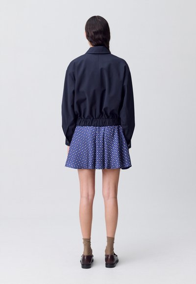 Chaqueta azul marino con cinturilla elástica, combinada con una falda acampanada que presenta un patrón de lunares azules y blancos. Calcetas y zapatos marrones.