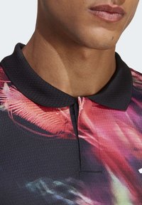 Polo a maniche corte con colletto nero, vivaci motivi floreali astratti in tonalità di rosa e rosso, e un tessuto liscio e texturizzato.