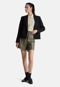 Czarna marynarka w prążki z złotymi guzikami, kremowa koszula, oliwkowozielone skórzane szorty, czarne teksturowane skarpetki oraz czarne buty z ozdobnym paskiem.