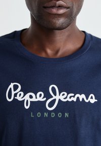 Tamsiai mėlynas medvilninis marškinėliai su baltais rankraščiu "Pepe Jeans" ir žaliais "LONDON" tekstais, trumpomis rankovėmis ir apvaliu kaklu.