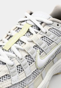 Svijetlosivi atletski tenisice s mrežastom i kožnom teksturom, bijelim vezicama, suptilnim žutim akcentima i istaknutim bijelim Swoosh logom.