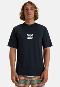 Mörkblå rash guard med kort ärm som är tillverkad av slätt, stretchigt material och har en vit våg-logotyp på bröstet. Kombinerad med simshorts i flerfärgade ränder.