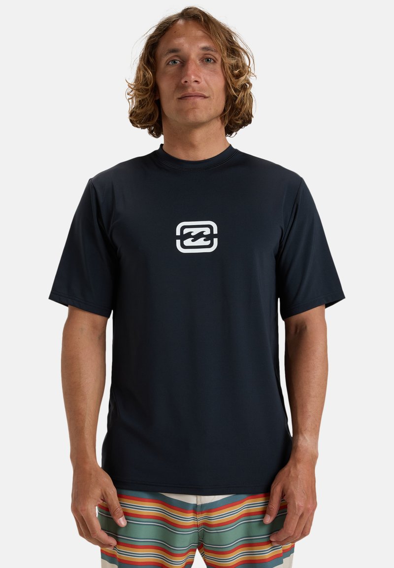 Mörkblå rash guard med kort ärm som är tillverkad av slätt, stretchigt material och har en vit våg-logotyp på bröstet. Kombinerad med simshorts i flerfärgade ränder.