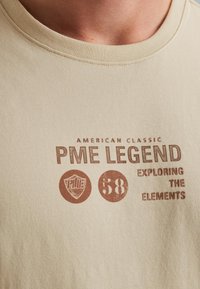 Béžové bavlnené tričko s okrúhlym výstrihom. Obsahuje potlačený text hnedou farbou: "PME LEGEND," štítový znak a číslo "58."