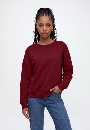 Donna con capelli intrecciati che indossa orecchini d'oro, una felpa rosso scuro e jeans blu, in piedi con una mano in tasca su sfondo bianco.