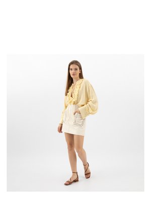 Femme marchant, portant un chemisier jaune pâle à volants, une jupe blanche avec des poches brodées et des sandales plates marron à brides sur un fond blanc.