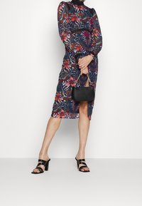 Šaty s květinovým vzorem a dlouhými rukávy, s vysokým límcem, v živých barvách na tmavém pozadí. Nosené s černými sandály na podpatku a malou kabelkou.