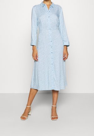 Woman wearing a light blue, long-sleeve, button-down midi dress with a small floral pattern and white heeled sandals.