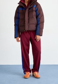 Bruine donsjas met blauwe strepen, gecombineerd met bordeauxrode trainingsbroek met blauwe accenten. Oranje schoenen maken de outfit compleet.