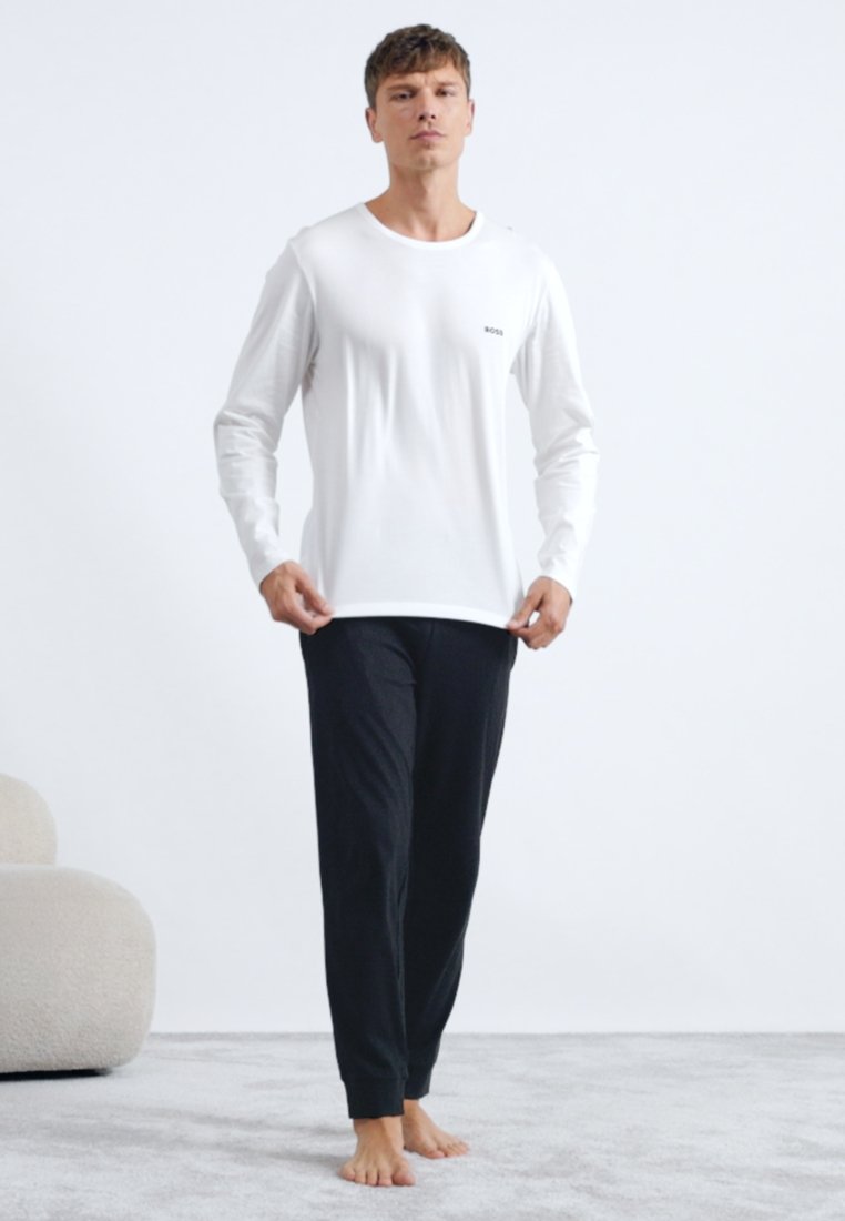 Chemise blanche à manches longues avec un petit logo noir, associée à un pantalon noir. Le matériau semble doux ; la coupe est décontractée.