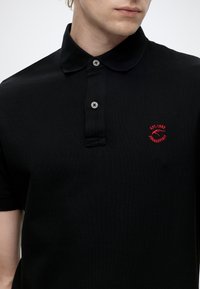 Young person wearing a black polo shirt with two buttons and a small red embroidered logo on the left chest.
