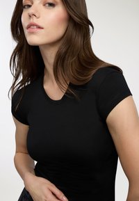 T-shirt nera aderente con maniche corte, colletto rotondo, texture liscia e dettagli di cucitura minimal, indossata da una persona con lunghi capelli castani.