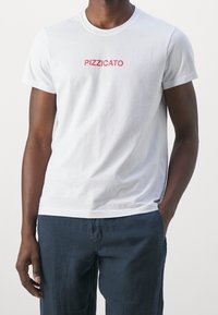 Hvid bomulds t-shirt med rød "PIZZICATO" print. Korte ærmer, rund halsudskæring og en afslappet pasform. Bæres med mørkeblå bukser.
