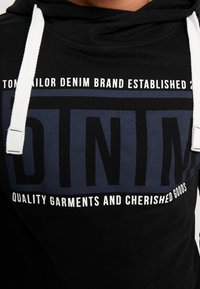 Zbliżenie czarnej bluzy z kapturem z białymi sznurkami, z napisem "Tom Tailor Denim Brand Established" oraz "Quality Garments and Cherished Goods."