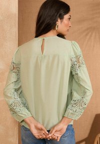 Blusa verde chiaro con maniche lunghe, caratterizzata da delicati dettagli in pizzo sulle maniche e un'apertura a chiave sulla schiena. Tessuto morbido e vestibilità rilassata.