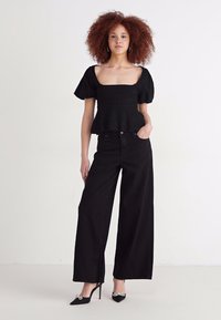 Haut peplum noir en tissu texturé avec manches bouffantes, associé à un pantalon large noir et des escarpins noirs à talons hauts, modèle présenté.